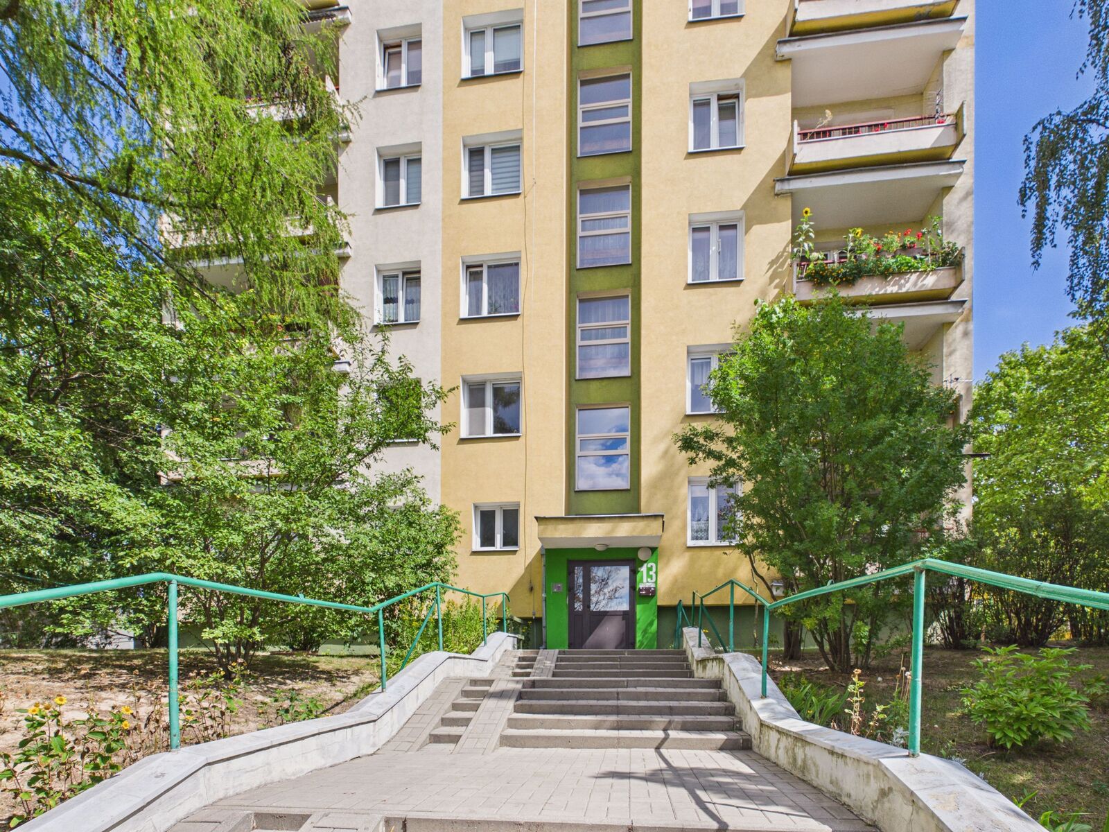 Wejście do żółtego bloku mieszkalnego otoczonego zielenią